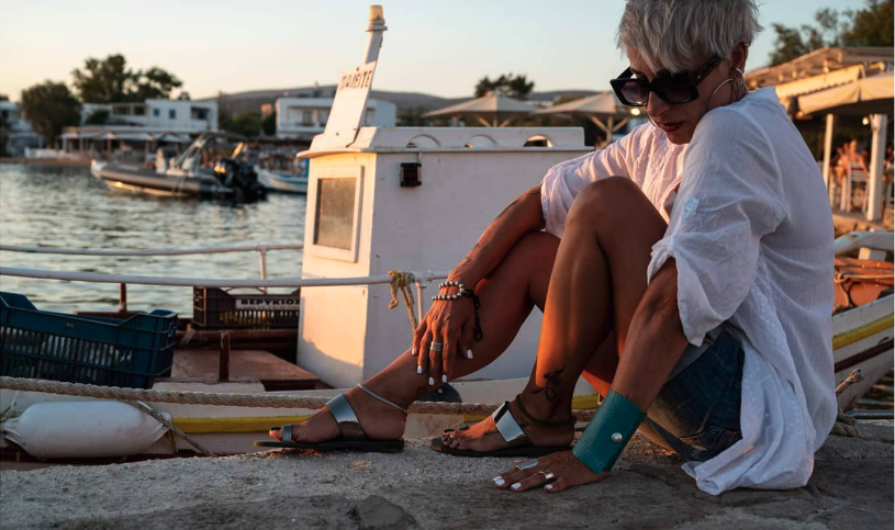 Woman sitting on the edge of a body of water at sunset, featuring bold jewelry she is wearing